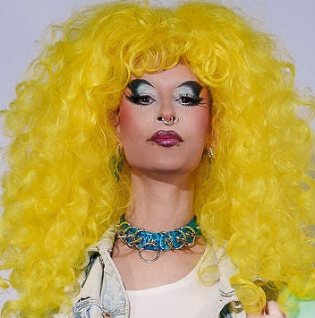 Person with bright yellow wig and colorful outfit holding a handbag against a plain background