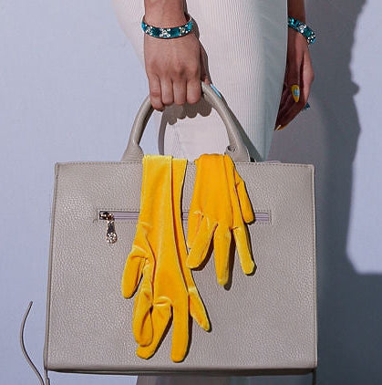 Person with bright yellow wig and gloves holding a handbag against a plain background