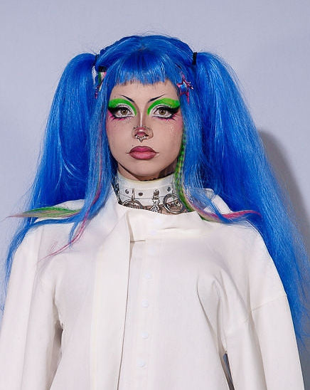 Person with blue hair and a white coat standing against a plain background