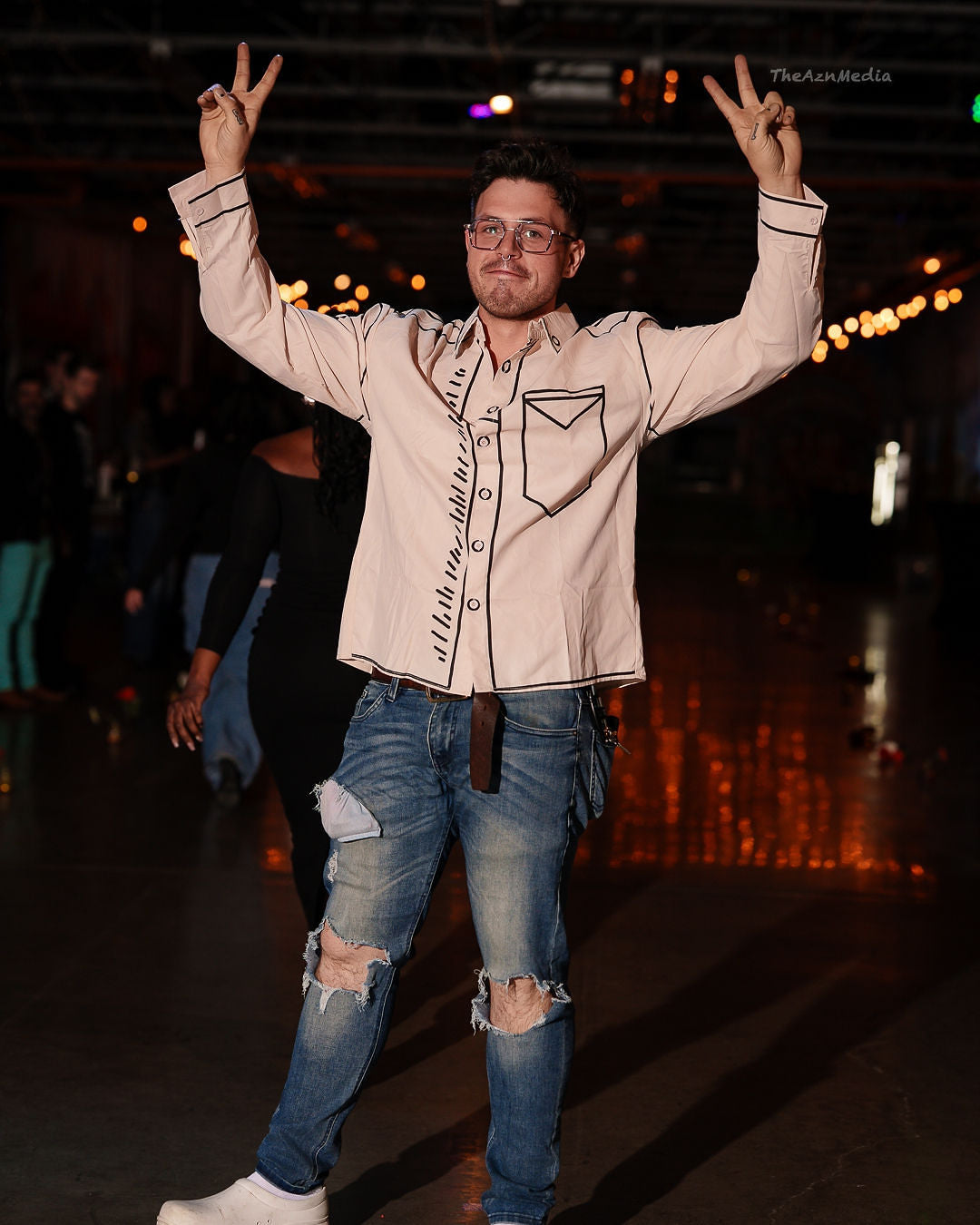 JACOB C. SCOTT in a beige shirt and ripped jeans making a peace sign gesture on a dark background.
